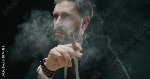 Pensive bearded male in elegant suit smoking cigar in dark room full of smoke, closeup photo