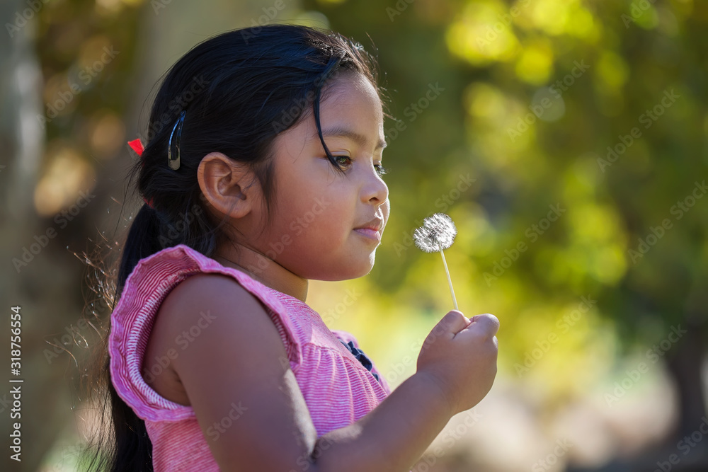 A thouthful little kid holding a dandelion in her hand that is close to her face and surrounded by green foliage. A thouthful little kid holding a dandelion in her hand that is close to her face and surrounded by green foliage.