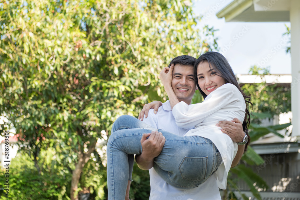 Young loving couple have fun in park. Caucasian boyfriend carry on his asian girlfriend in arms in front yard of house.