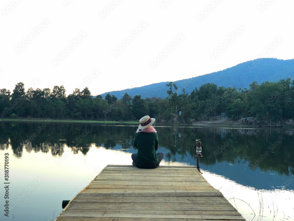 Rear view on young woman sitting on wooden pier in the evening looking at calm lake