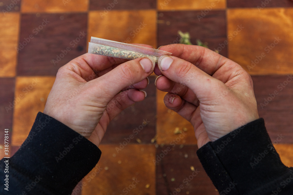Fototapeta premium Overhead closeup shot of fingers pinching the end of a ground cannabis filled rolling paper forming it into a joint over a checkered table in the background.
