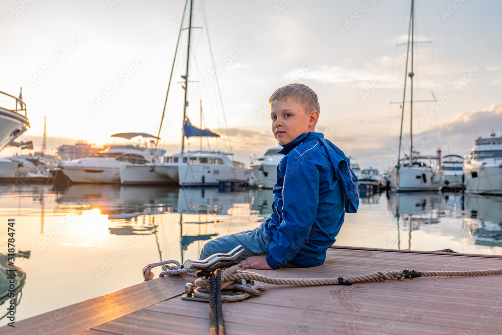 A boy in a blue jacket sits on a pier with yachts during sunset.