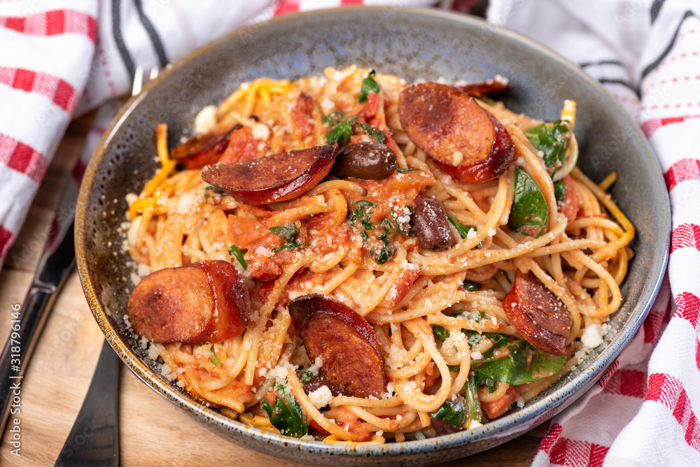 A bowl of chef prepared spaghetti with chorizo, black olives, and spinach in a creamy tomato sauce in a ceramic bowl on a wooden chopping board with red and white napery. A bowl of chef prepared spaghetti with chorizo, black olives, and spinach in a creamy tomato sauce in a ceramic bowl on a wooden chopping board with red and white napery.