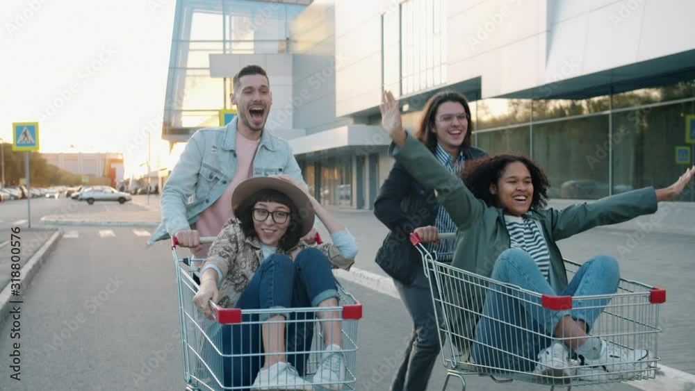Students young men and women are riding shopping carts outside doing high-five laughing together. Happiness, entertainment and friendship concept.