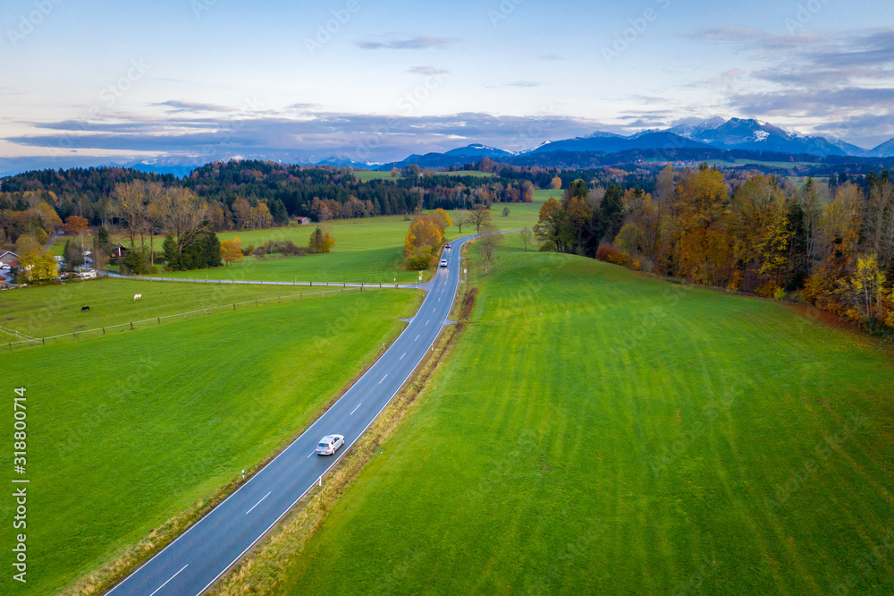 Aerial view Autumn landscape in Bavaria near Miesbach. Road going into the distance and the peaks of the Alps on the horizon. Clouds sky, Yellow trees and green field
