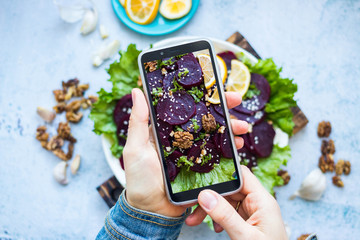 Woman take picture of vegan food with phone at her kitchen. Hand make a closeup smartphone photo of beetroot salad with walnuts for blogging or social media content. Vegetarian healthy food.