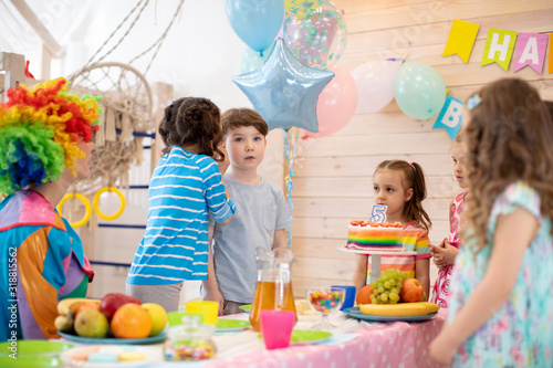 Group of adorable kids standing around festive table at party. Little girl congratulating birthday boy.