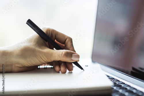Closeup of woman hand writing in diary on a table with laptop. Focused on a pen