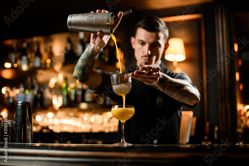 Male bartender pouring a smooth yellow alcoholic cocktail drink from the steel shaker through the sieve