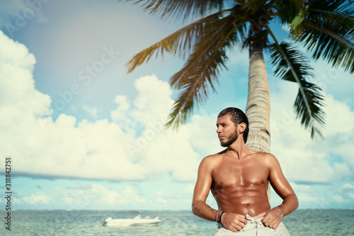 Enjoying suntan and vacation. Portrait of young bearded man leaning on coconut palm tree on the tropical beach.