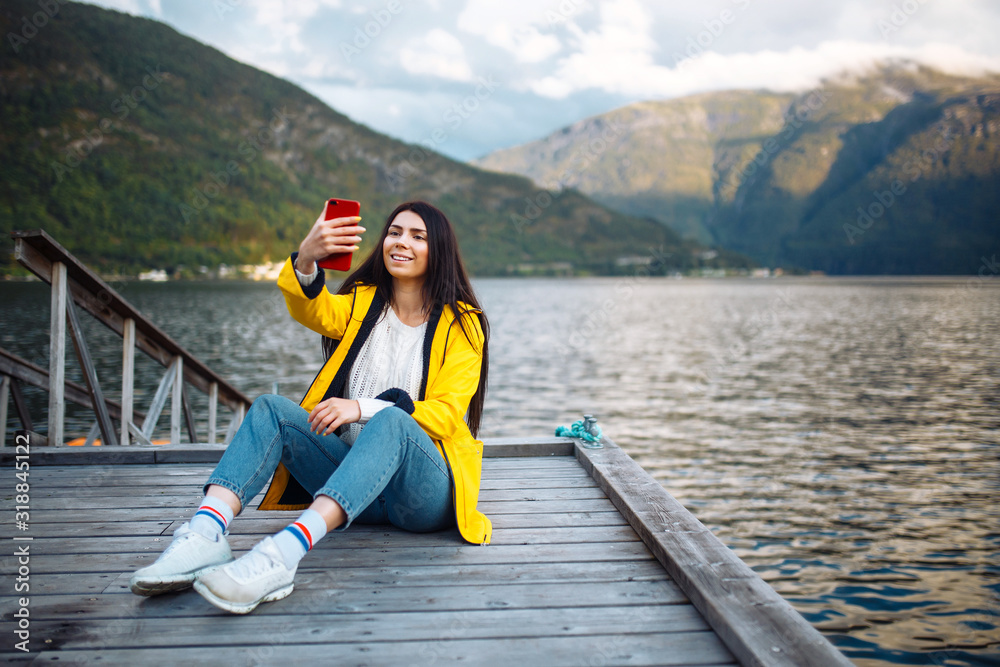 The girl tourist takes a photo on the phone by the lake in Norway. Young woman takes selfie against the backdrop of the mountains in the Norway. Travelling, lifestyle, adventure, concept. The girl tourist takes a photo on the phone by the lake in Norway. Young woman takes selfie against the backdrop of the mountains in the Norway. Travelling, lifestyle, adventure, concept.