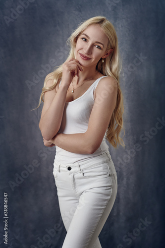 Attractive sexy slender blond woman with long hair posing in off white summer top and jeans with her hand to her chin smiling at the camera over a grey studio background in a three quarter portrait