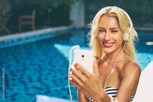Music and vacation. Enjoying summertime. Pretty young woman in bikini using smartphone in headphones near swimming pool.