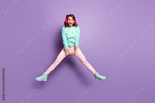 Full length photo of pretty lady jumping high up excited good mood spread legs sides having fun wear fuzzy pullover pink ear muffs pastel pants shoes isolated purple color background