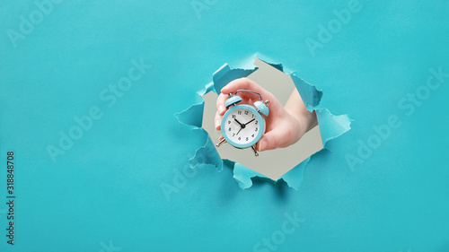 Hand holds alarm clock through a paper hole. Time management and deadline concept
