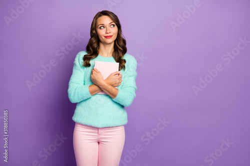 Photo of beautiful curly lady hold favorite love novel close to chest look empty space dreamer thinking what to read next wear pastel fuzzy sweater pink pants isolated purple color background