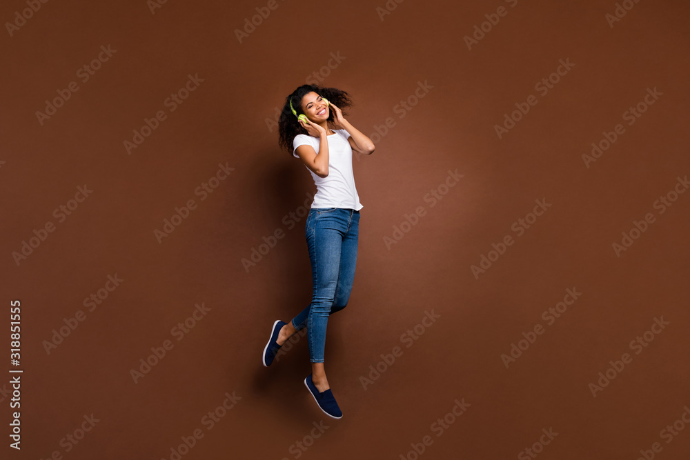 Full size photo of cheerful afro american girl have free time listen playlist music use green headset enjoy stereo sound jump wear stylish white t-shirt isolated over brown color background Full size photo of cheerful afro american girl have free time listen playlist music use green headset enjoy stereo sound jump wear stylish white t-shirt isolated over brown color background