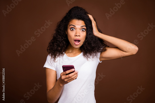 Portrait of astonished afro american girl blogger use cell phone read information stare stupor hand touch head scream omg wear casual style outfit isolated over brown color background