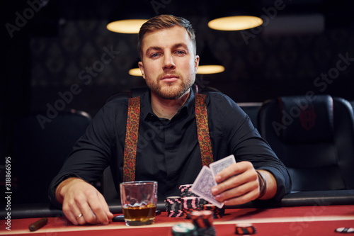 Elegant young man with glass of alcohol sits in casino and plays poker game