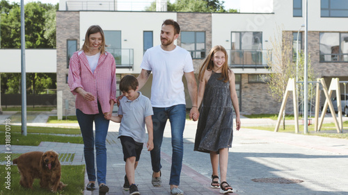 Happy affectionate young family walking in the neighbourhood of their new house. Smiling parents and their children holding hands exploring the new place on a sunny day.