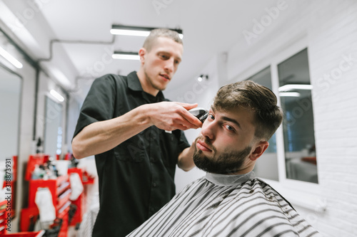 A fun client cuts a professional male hairdresser, sits on a hairdresser and looks into the camera. Barber cuts a cheerful bearded client. Barber shop concept.