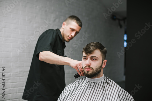 Professional barber cuts a bearded client in a barbershop, looks at her hair and uses a trimmer. Positive male hairdresser makes male haircut with a clipper. Barbershop concept