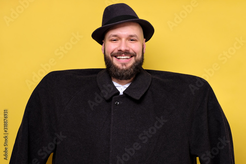 Man in oversize baggy black coat and hat smiling standing on yellow studio