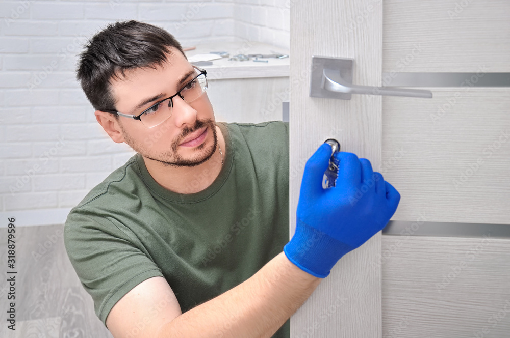 The guy installs new handles and a lock for the door to the room. Repair in the house. The guy in the blue gloves holds the door handles. Installing a door lock. - obrazy, fototapety, plakaty 