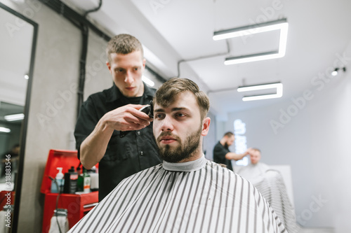 Barber haircuts a handsome hairy man in a light men's hairdresser, the client looks forward, the hairdresser is working on a hair clipper. Men's haircuts in barbershop concept.
