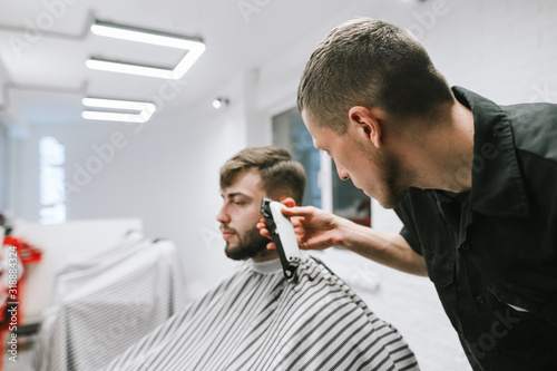 Professional male hairdresser cuts bearded man in modern barbershop, uses clipper. Focusing on a barber with a clipper creates a stylish hairstyle for the client.