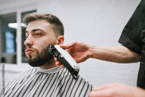 Barber trims the beard of a handsome man with a clipper. Closeup portrait of barber shop client. Barber correction of client's beard in men's hairdresser.