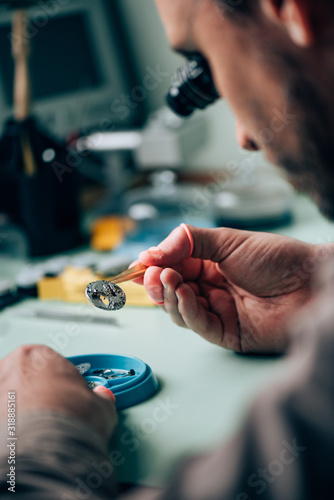 Selective focus of clockmaker in eyeglass loupe holding watch part in tweezers by tool tray on table