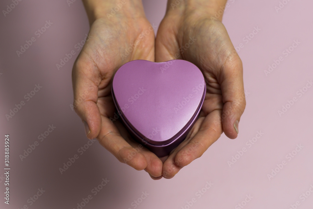 Fototapeta premium In female young hands lies a small heart-shaped box, close-up, on a light pinkish background.