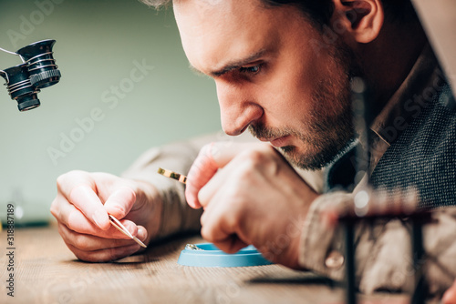 Side view of handsome watchmaker using magnifying glass for clock part at working table photo
