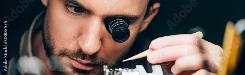 Panoramic shot of watchmaker in eyeglass loupe working with wristwatch isolated on black photo