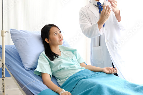 Young male Asian doctor in medic uniform holding syringe in hand at hospital with Anxiety female Asian patient lie in bed and looking at the doctor, Healthcare and medically concept