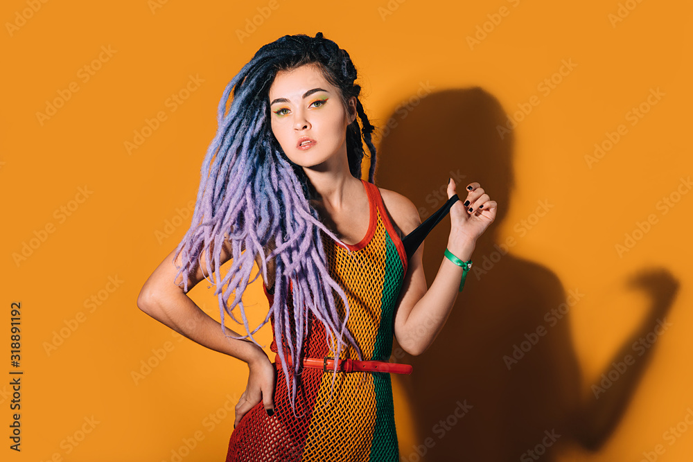 Fotka „Hipster woman with violet dreadlocks posing on camera. Rastafarian  woman in a rasta dress on orange background“ ze služby Stock | Adobe Stock