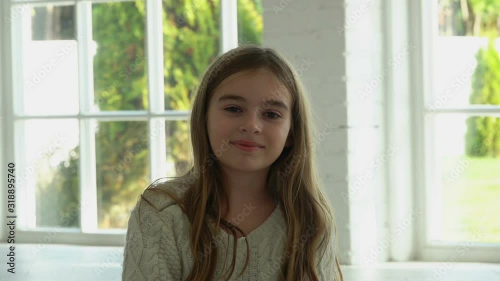 Cute child girl with a long hair is smiling and sitting in a white sunlit hall with big windows Cute child girl with a long hair is smiling and sitting in a white sunlit hall with big windows