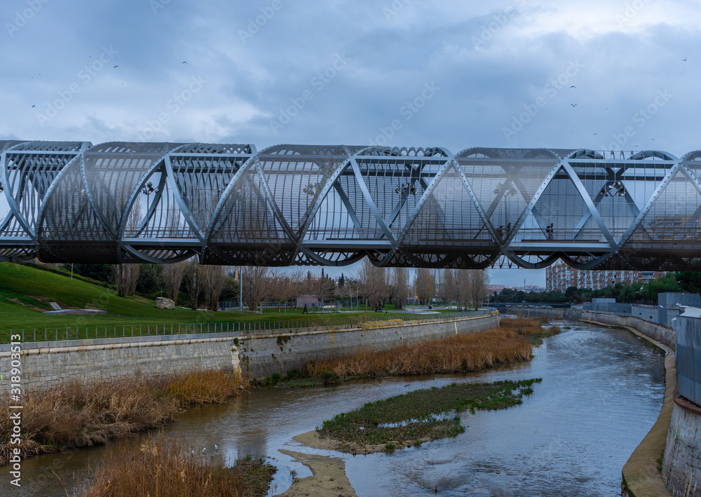 Obraz premium Views of the Arganzuela Bridge on a cloudy day, pedestrian bridge crossing the Manzanares River in the Madrid Río Park, formed by a metal spiral covered with a mesh, travel concept