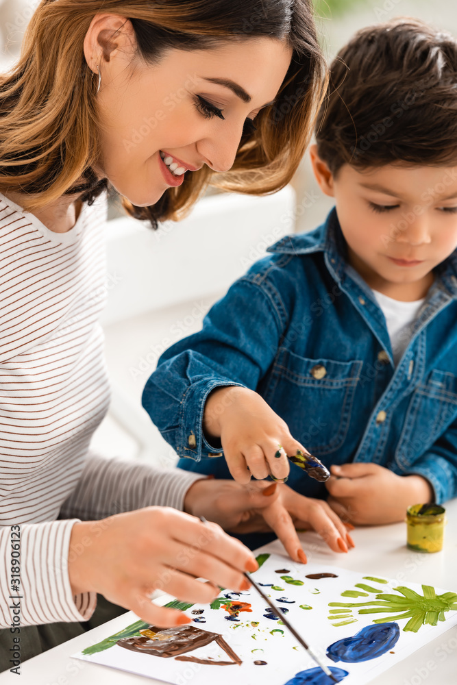 Fototapeta premium happy woman sitting at table and drawing with paints together with son