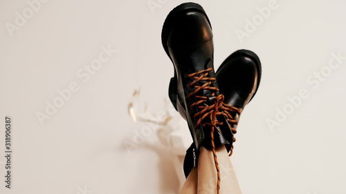 The naked woman legs wearing only black leather boots upside down in front of a white wall. Black fashion shoes with red shoelaces upside down.