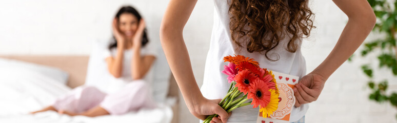 back view of daughter holding flowers and mothers day card while mom sitting in bed, panoramic shot