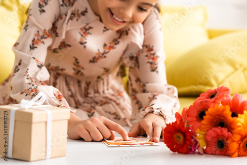 selective focus of cute child making mothers day card while sitting near flowers and gift box
