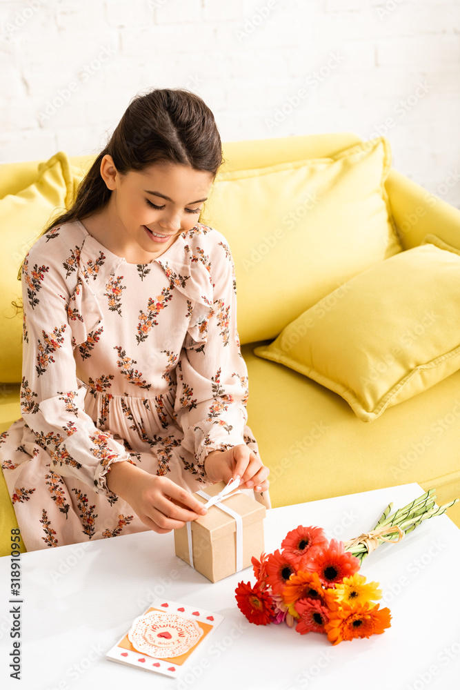 smiling child in elegant dress tying ribbon on gift box while sitting near flowers and mothers day card smiling child in elegant dress tying ribbon on gift box while sitting near flowers and mothers day card