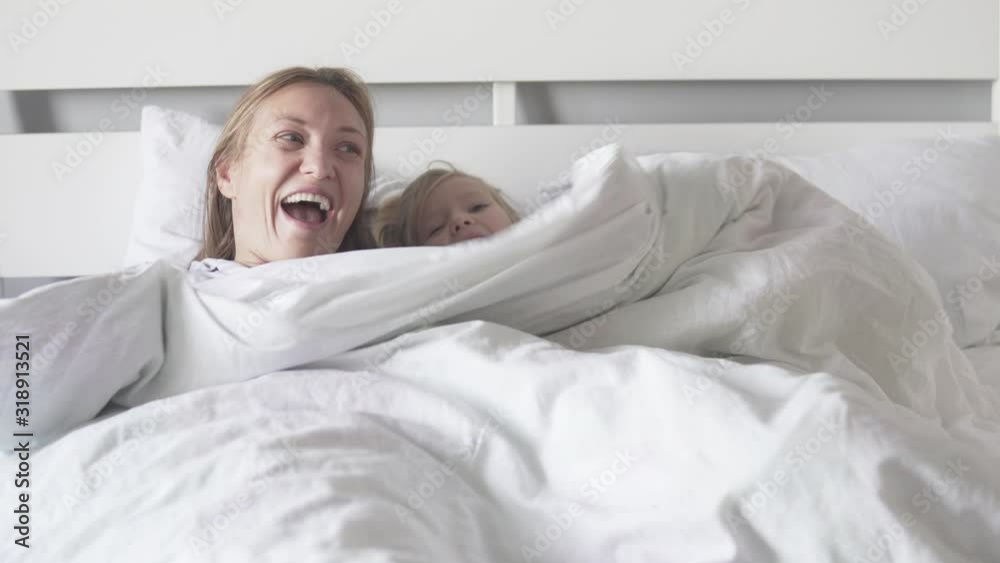Mom and daughter hide under a blanket in the bed. Mom and baby peek out from under blankets and laugh cheerfully Mom and daughter hide under a blanket in the bed. Mom and baby peek out from under blankets and laugh cheerfully