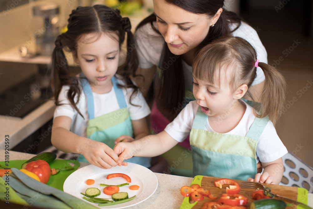 happy family mom and her little daughters are preparing healthy food, they make smiley face with vegetables morsels in domestic kitchen happy family mom and her little daughters are preparing healthy food, they make smiley face with vegetables morsels in domestic kitchen