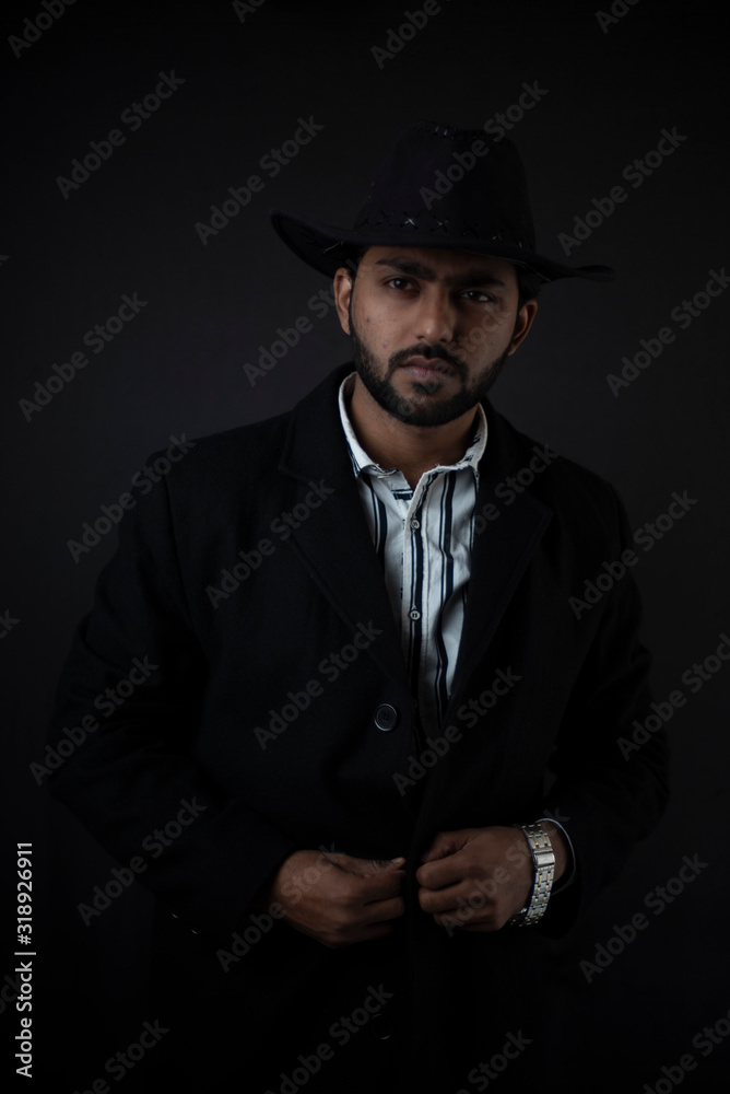 Fashion portrait of an young and handsome Indian Bengali brunette man with striped formal shirt, black suit and cowboy hat in black copy space background. Indian lifestyle and fashion photography.