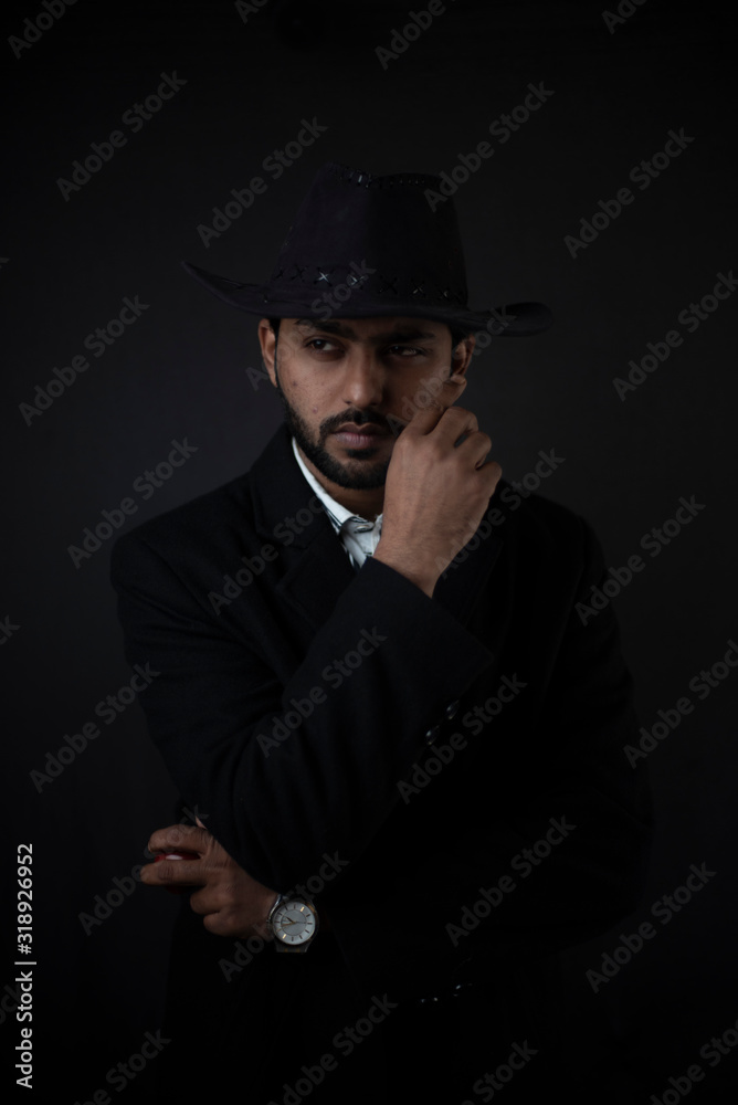 Fashion portrait of an young and handsome Indian Bengali brunette man with striped formal shirt, black suit and cowboy hat in black copy space background. Indian lifestyle and fashion photography.