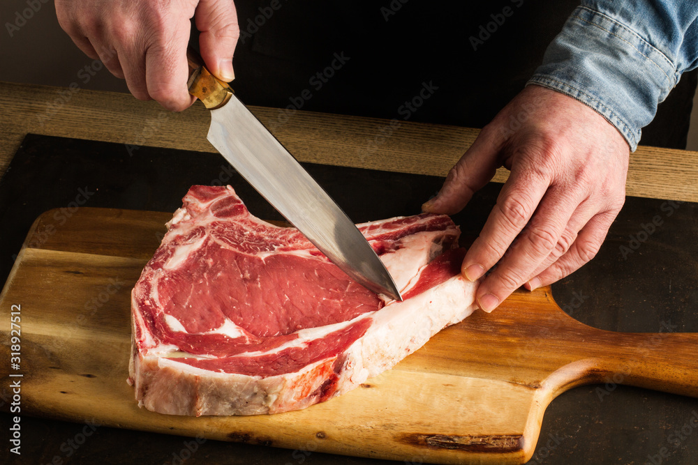 Hombre con cuchilla en mano cortando preparando chuletón de vaca sobre tabla de madera rústica. Vista de frente y superior