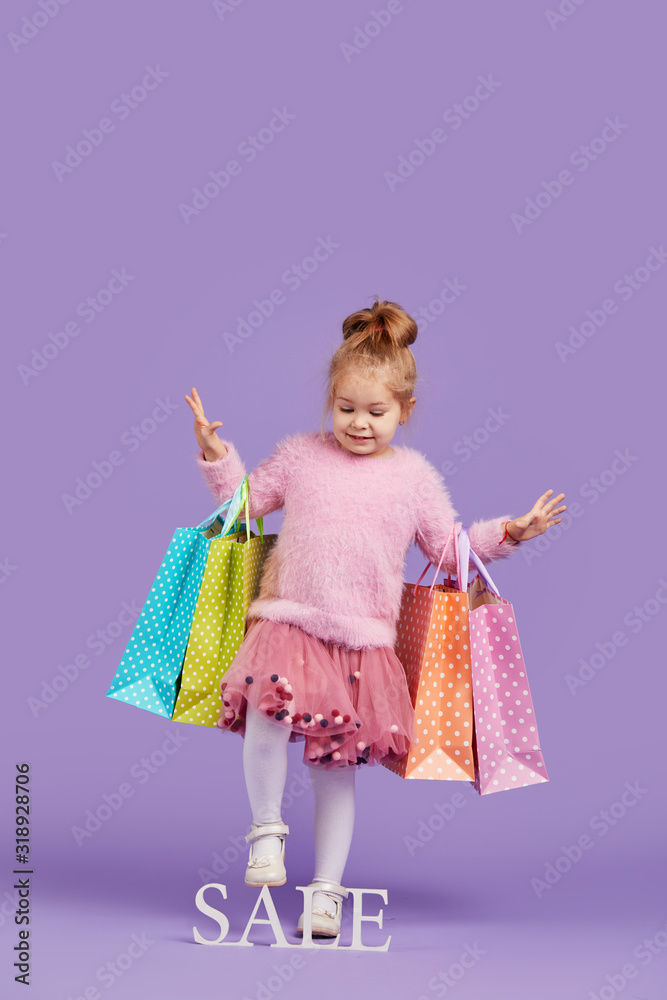 Portrait of a happy little child girl on a purple background holding shopping bags, package. Looking at the camera. Sale concept
